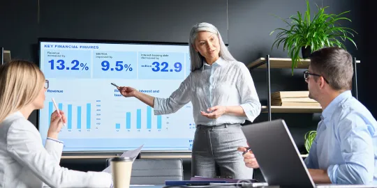 A performance marketer presenting financial data on a screen in front of two people in a meeting room.